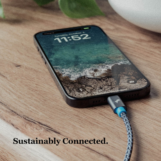 Banner image with iPhone connected to black and white powerknit cable with blue and gray cable head on wood surface. Text overlay Sustainably Connected.