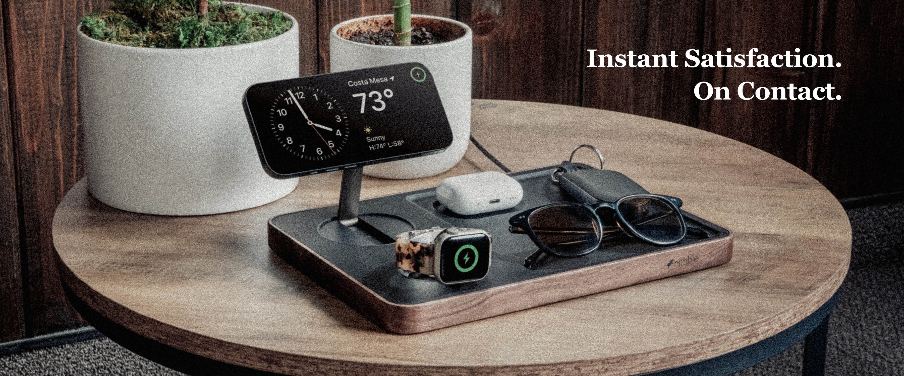 Banner with Valet 3-in-1 Wirelss Charger with iPhone, AirPods, Apple Watch charging and Glasses, keys placed as well. All is on small wood table with potted bamboo plants and wood wall. Text overlay Instant Satisfaction. On Contact.