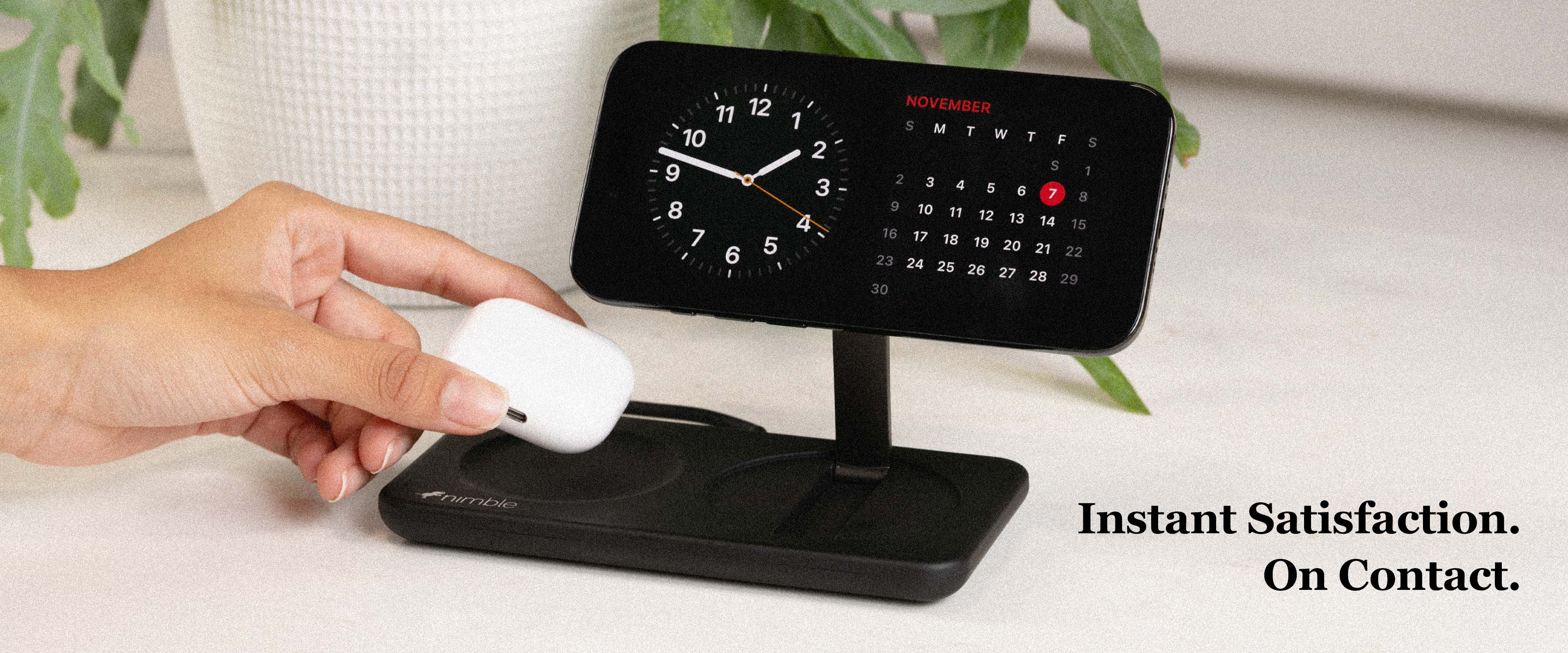 Black Podium 2-in-1 Wireless Charger  on white kitchen surface with iPhone charging and hand placing AirPods to charge. Text overlay Instant Satisfaction. On Contact.