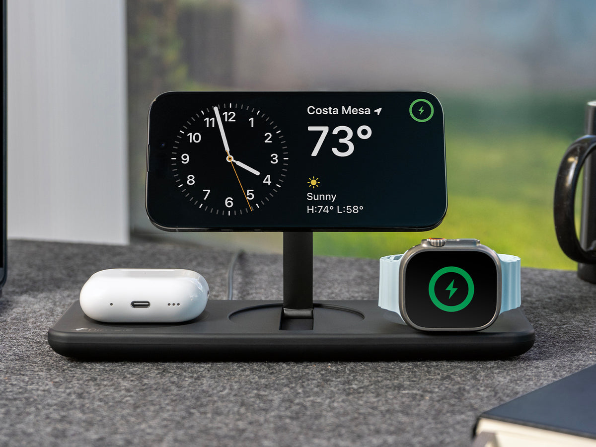 Podium 3-in-1 25W Wireless charging stand with iPhone, AirPods and Apple Watch devices charging upright on a felt desk surgace with a blurred outdoor background
