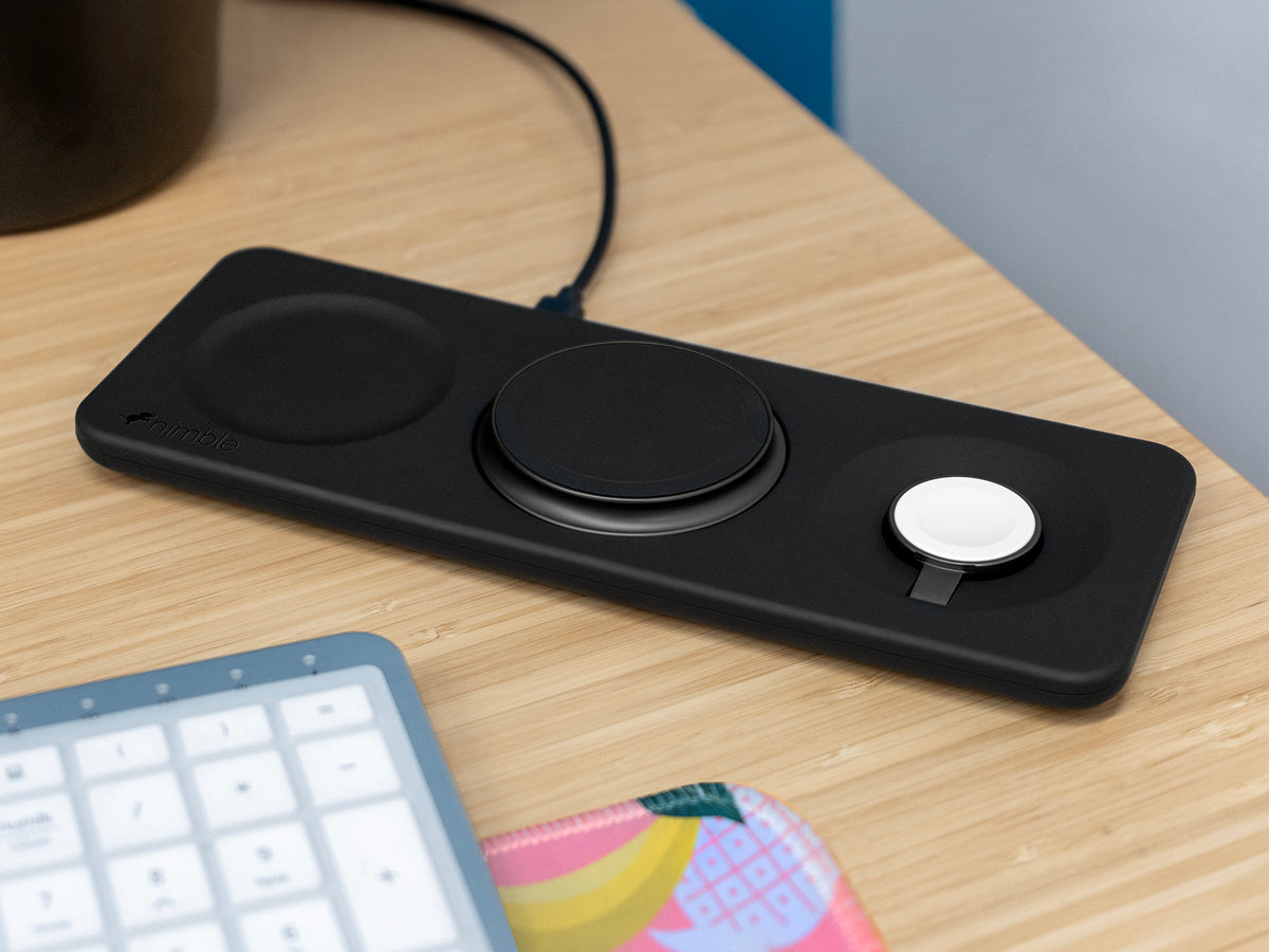 Podium 3-in-1 25W Wireless charging stand with dedicated spaces to charge iPhone, AirPods and Apple Watch devices on a desk with a keyboard and colorful mouse pad.