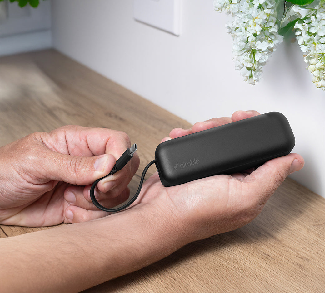 Person holding a black Wally Pro 20 wall charger with built-in battery with USB-C cable, on a wooden surface with outlet and flowers in the background.