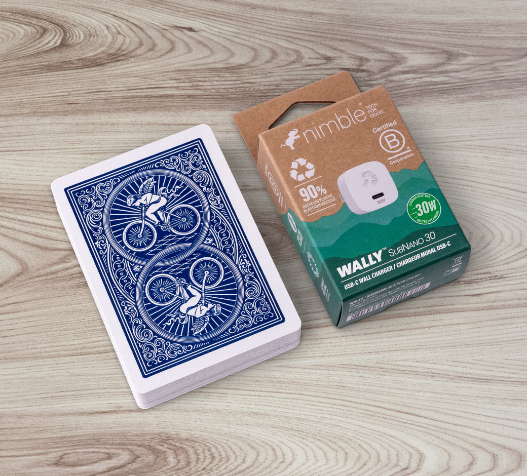 A deck of playing cards with a blue Bicycle design next to a Nimble WALLY SUBNANO 30 USB-C wall charger in a cardboard box on a wooden surface.
