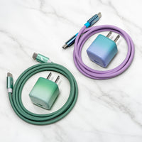 Two coiled USB-C charging cables in Coastal Moss/Emerald Cove with matching wall chargers are arranged on a white marble surface. One set is mint green to teal, and the other is lavender to blue.